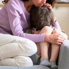 En mamma tröstar sitt barn som kramar sina knän. Barnets ansikte är dolt, men kroppsspråket visar oro.