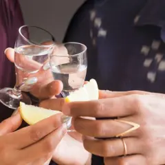 Två personer skålar med små glas med klar alkohol. Citronklyftor och salt ligger redo för att förhöja smaken av gaba alkohol.