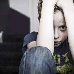 En pojke sitter på en trappa med händerna över huvudet, blicken tom. Bilden symboliserar känslan av obehandlad PTSD.