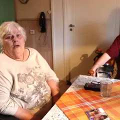 En kvinna med vitt hår sitter vid ett bord. Hon ser trött ut, som om hon inte orkar med sin dementa mamma. En man sitter mittemot.