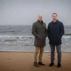 Två män står på en strand. Vågorna rullar in mot land. Det känns svårt att prata i den här blåsten.