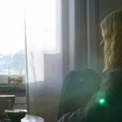 En kvinna sitter ensam vid ett fönster, med en kopp kaffe. Hon verkar drabbad av depression, kanske förvärrad av alkohol.