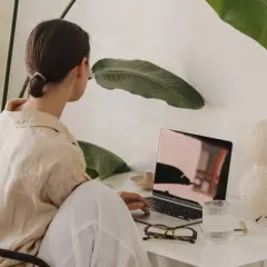 Kvinna arbetar vid laptop, omgiven av gröna växter. Kanske hon söker verktyg för att hantera kbt stress.
