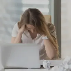 En kvinna med utmattningssyndrom känner yrsel och håller huvudet i händerna vid ett skrivbord med en laptop och papper.