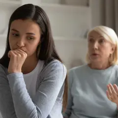 En ung kvinna sitter med handen mot munnen, ser bekymrad ut. Bakom henne gestikulerar en äldre kvinna, kanske en diskussion om ett problem med vuxen dotter.