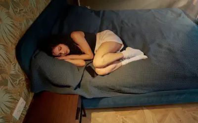 En ung kvinna sover hopkrupen på en säng, med en telefon bredvid sig. Hon verkar kämpa med **ångest** och **hyperventilera** i sömnen.