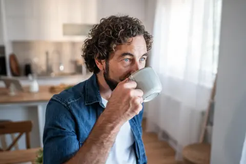 Man med lockigt hår dricker kaffe, blicken fundersam. Kanske en stund av kaffe ångest innan dagen börjar.