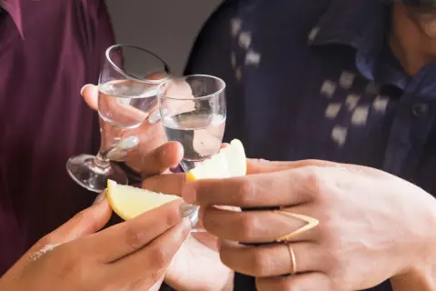 Två personer skålar med små glas med klar alkohol. Citronklyftor och salt ligger redo för att förhöja smaken av gaba alkohol.