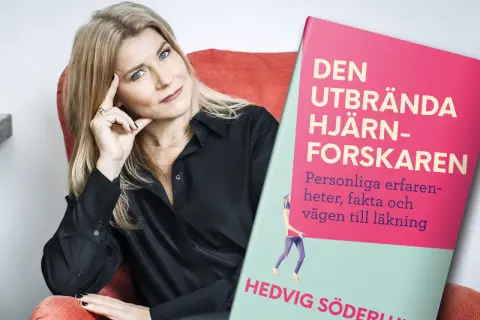 En kvinna med blå ögon och blont hår sitter i en röd fåtölj bredvid boken "Den utbrända hjärnforskaren". Boken handlar om utmattningssyndrom och hur man kan läka.