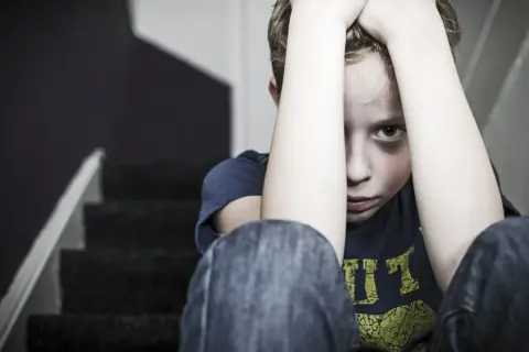 En pojke sitter på en trappa med händerna över huvudet, blicken tom. Bilden symboliserar känslan av obehandlad PTSD.