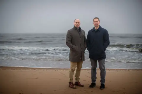 Två män står på en strand. Vågorna rullar in mot land. Det känns svårt att prata i den här blåsten.