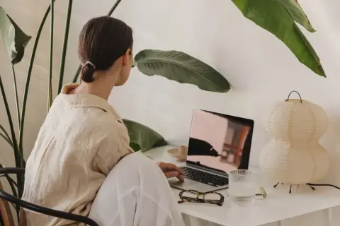 Kvinna arbetar vid laptop, omgiven av gröna växter. Kanske hon söker verktyg för att hantera kbt stress.