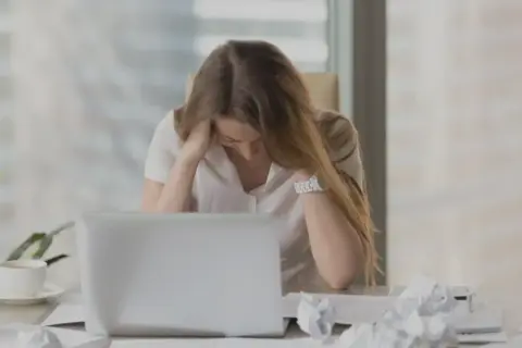 En kvinna med utmattningssyndrom känner yrsel och håller huvudet i händerna vid ett skrivbord med en laptop och papper.