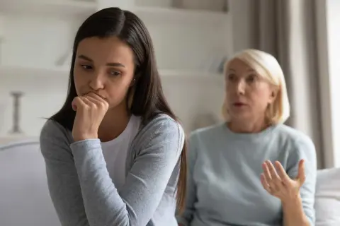 En ung kvinna sitter med handen mot munnen, ser bekymrad ut. Bakom henne gestikulerar en äldre kvinna, kanske en diskussion om ett problem med vuxen dotter.
