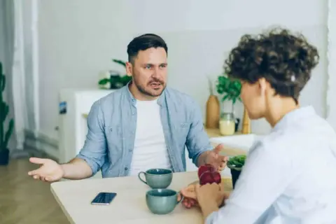 Man med intensiv blick förklarar något för kvinna, kanske om vad betyder narcissist. De sitter vid ett bord med kaffekoppar.