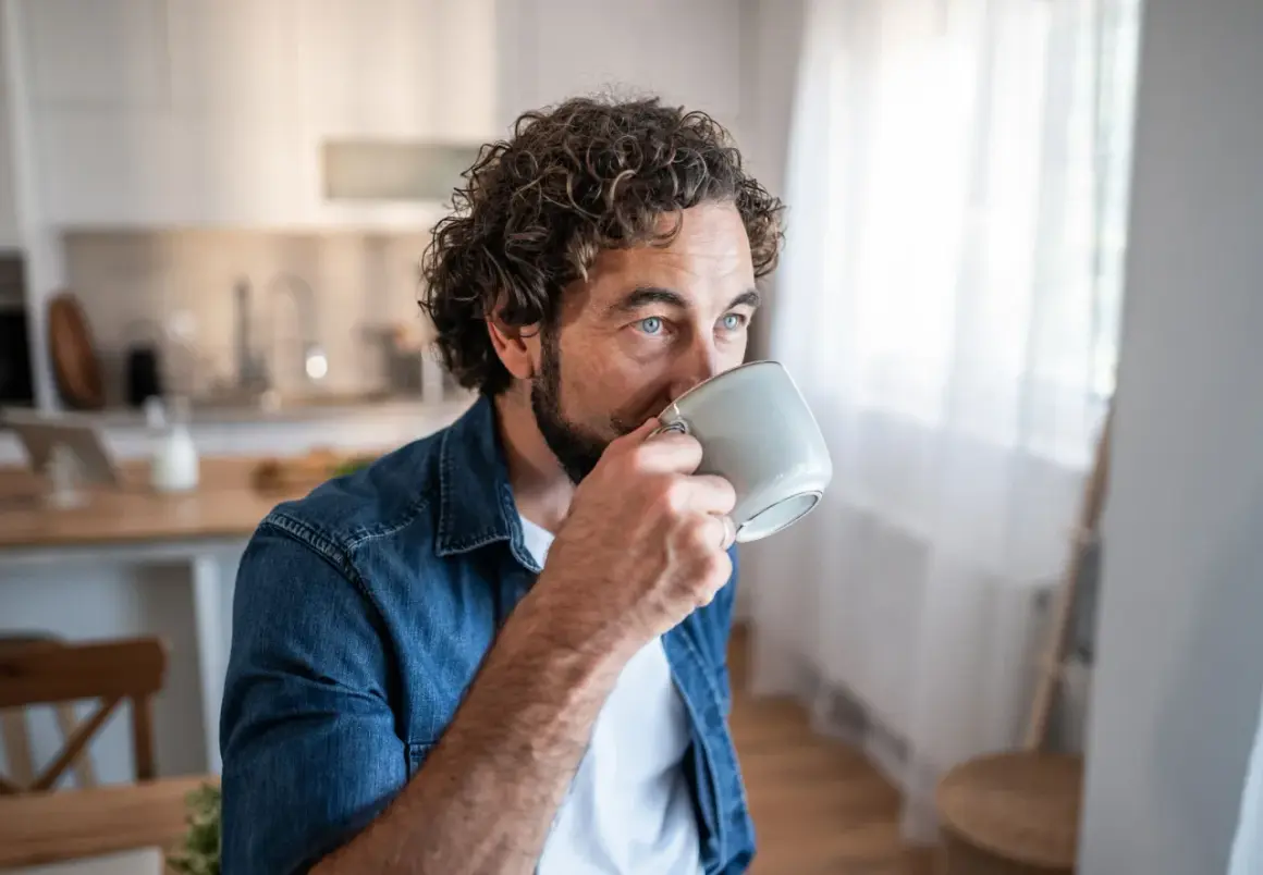 Man med lockigt hår dricker kaffe, blicken fundersam. Kanske en stund av kaffe ångest innan dagen börjar.