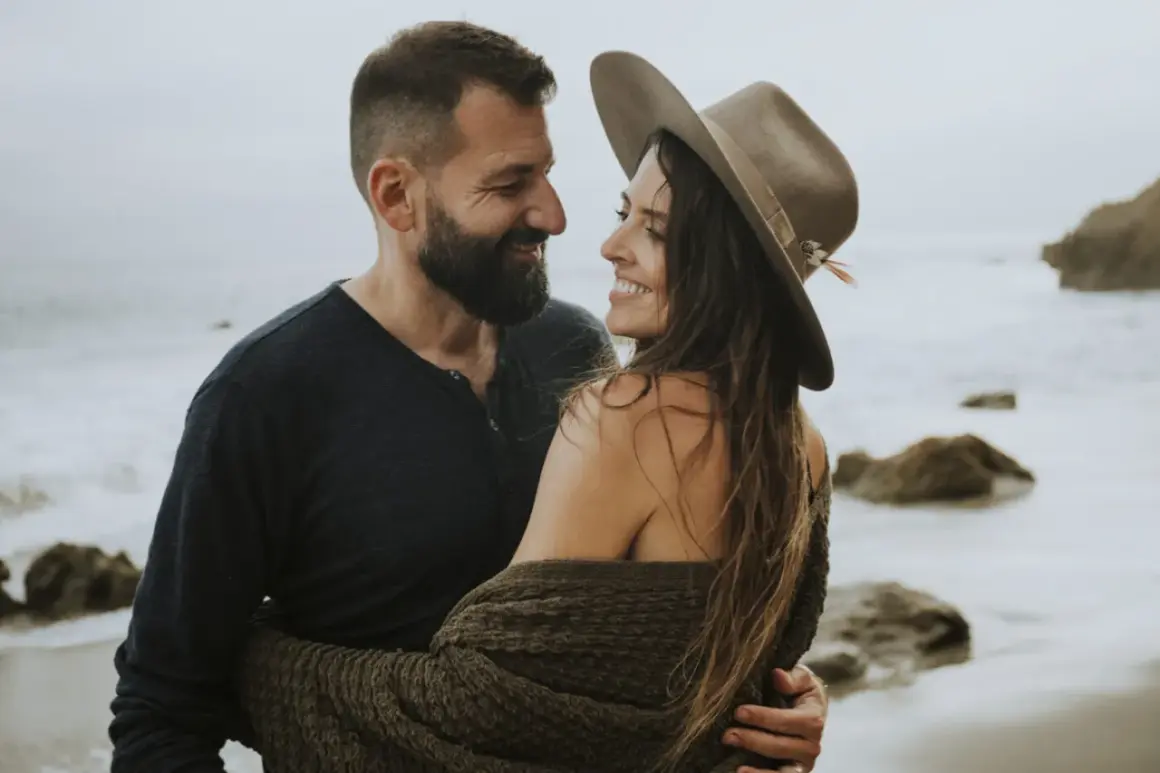 Ett par omfamnar varandra vid havet, deras blickar möts i en tydlig attraktion mellan två personer.