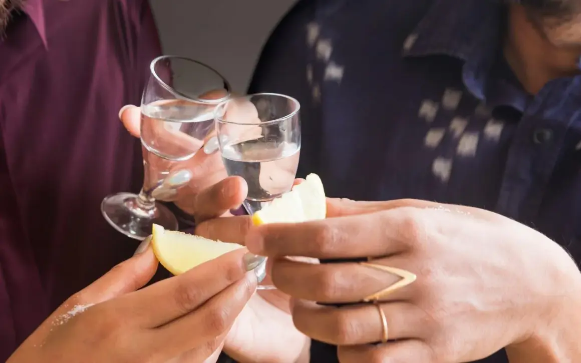 Två personer skålar med små glas med klar alkohol. Citronklyftor och salt ligger redo för att förhöja smaken av gaba alkohol.