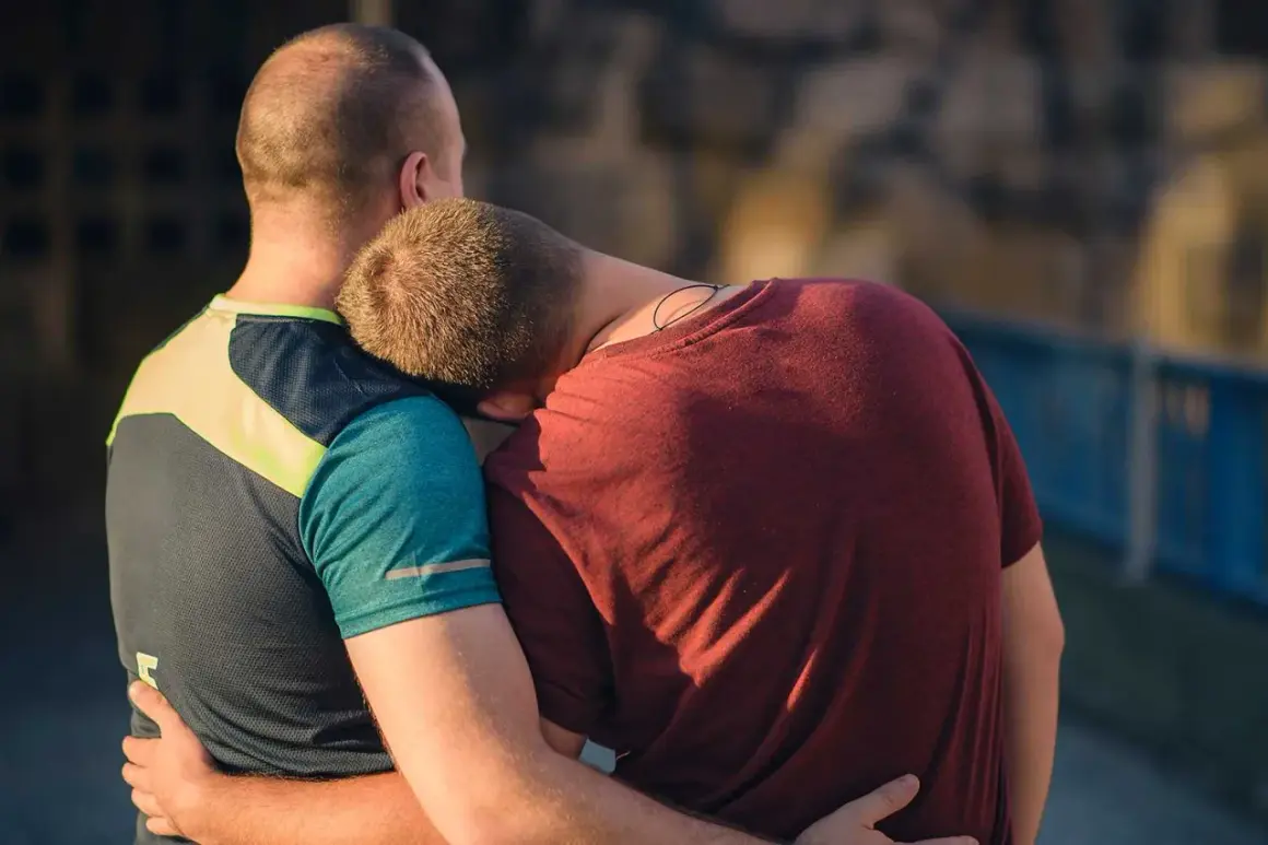 Två män omfamnar varandra. Den ena vilar huvudet mot den andras axel, en bild av en varaktig relation.