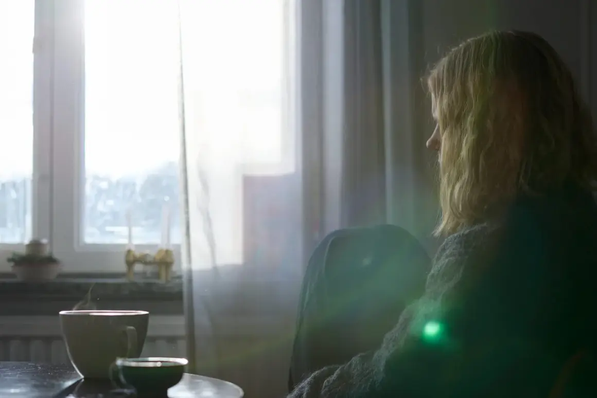 En kvinna sitter ensam vid ett fönster, med en kopp kaffe. Hon verkar drabbad av depression, kanske förvärrad av alkohol.