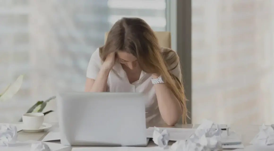 En kvinna med utmattningssyndrom känner yrsel och håller huvudet i händerna vid ett skrivbord med en laptop och papper.