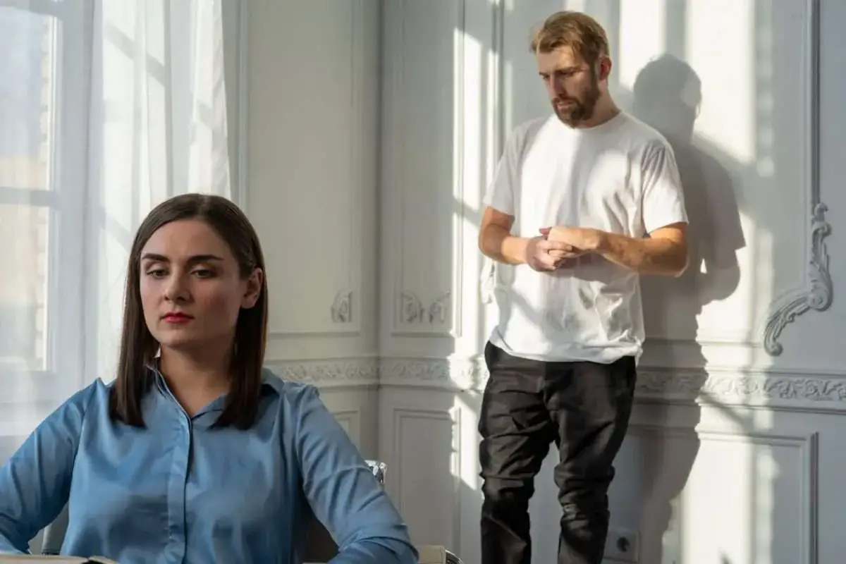 En kvinna med m&ouml;rkt h&aring;r sitter med blicken s&auml;nkt, medan en man med ljust sk&auml;gg st&aring;r bakom henne, med h&auml;nderna kn&auml;ppta. En k&auml;nsla av tystnad och ansp&auml;nning i deras medberoende relation.