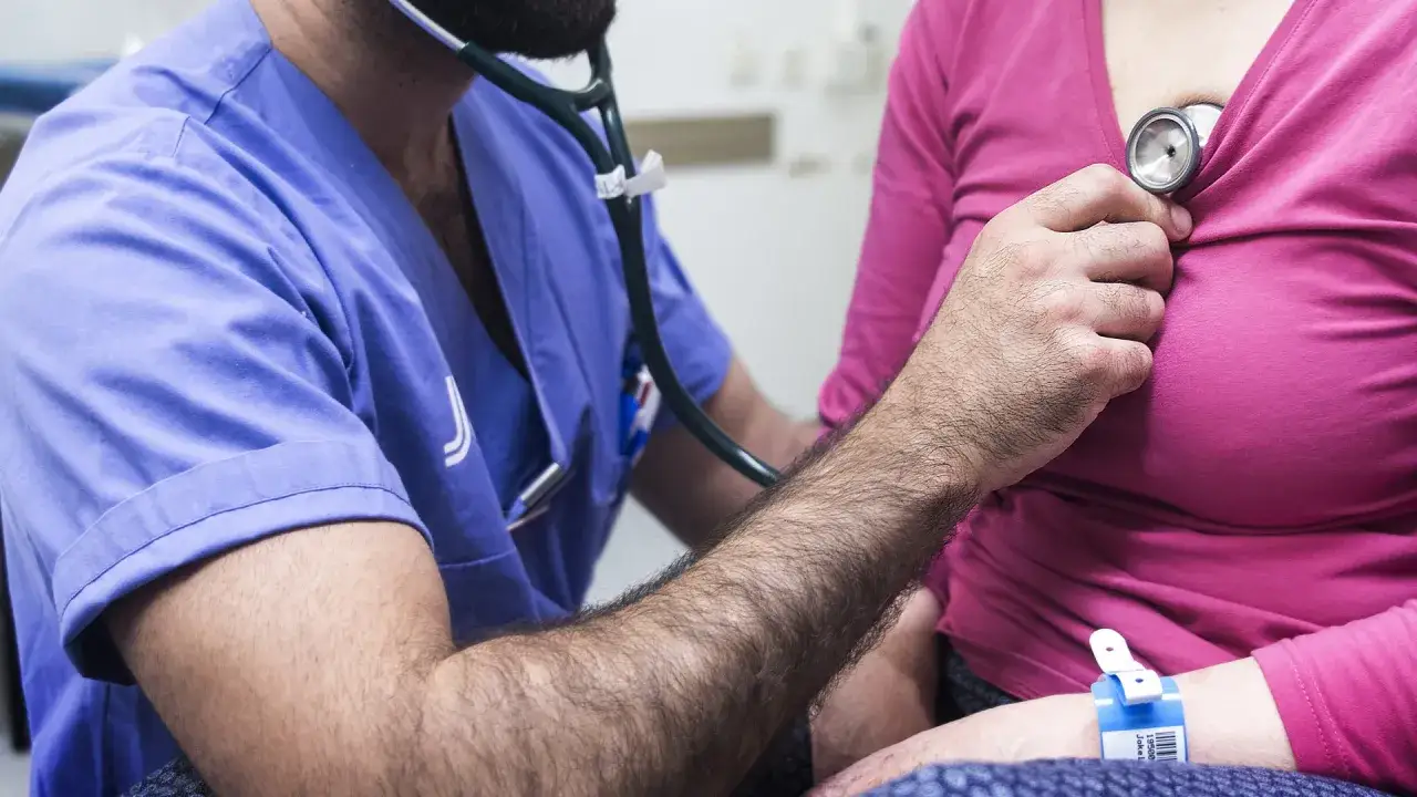 Läkare lyssnar på hjärtat med stetoskop. Patienten har ett armband, kanske för att få sjukskrivning vid utmattningssyndrom.