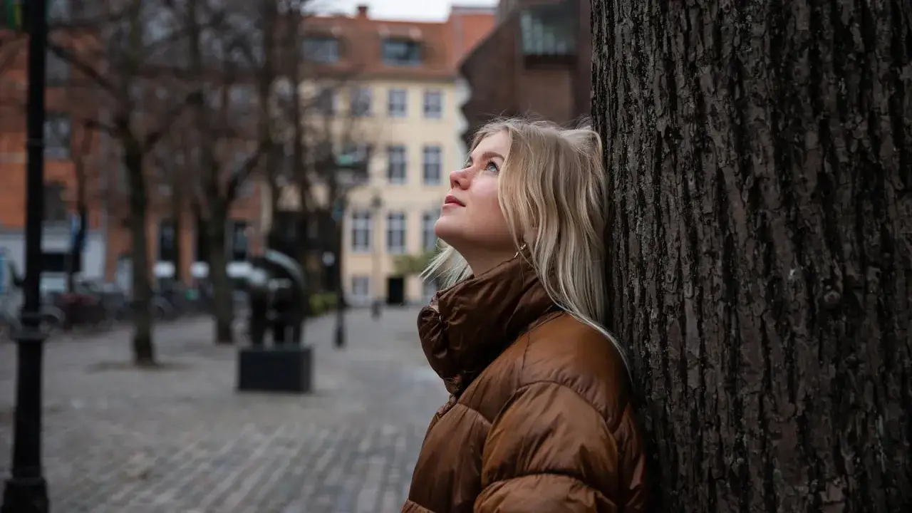 En ung kvinna lutar sig mot ett tr&auml;d, blicken upp&aring;t, kanske f&aring;ngad i sina &aring;ngest tankar. Stadsmilj&ouml; i bakgrunden.