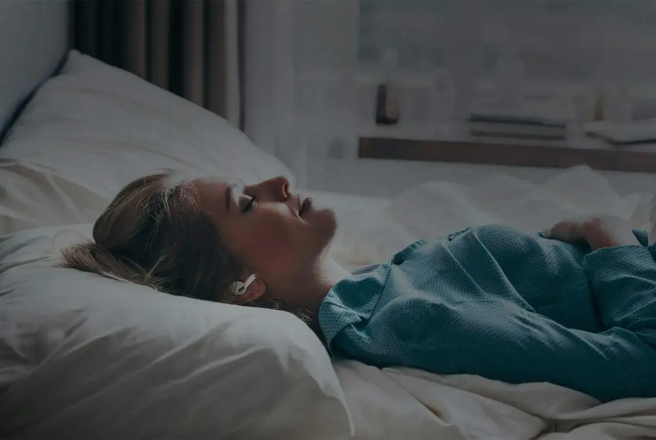 En kvinna ligger i s&auml;ngen med h&ouml;rlurar, ansiktet v&auml;nt upp&aring;t, kanske upplever &aring;ngest p&aring; morgonen.