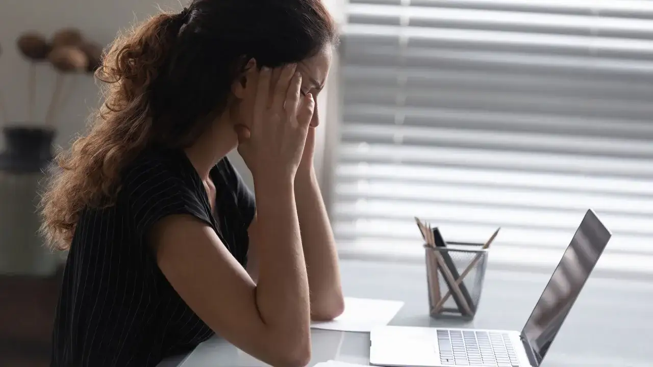 Kvinna med händerna mot tinningarna vid en laptop, uttrycker stress. Kanske dags för sjukskrivning.